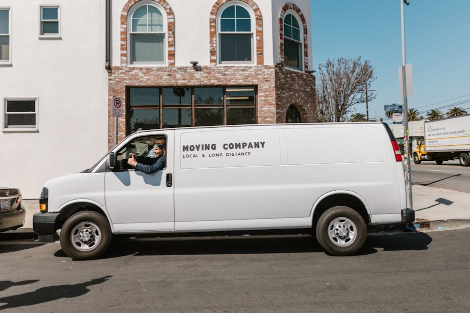 A side view of a white cargo van parked parallel to the curb on an urban street at night, with a dark building façade in the background. The van is positioned just off the pavement, with no visible d