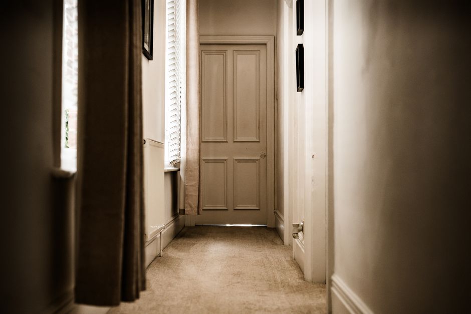 A narrow hallway inside a house, featuring beige walls and cream-colored carpet flooring. On the left side, a window with partially open white blinds allows natural light to enter, illuminating the sp