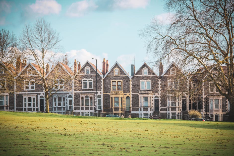 A row of five Victorian-style terraced houses built with dark brick and featuring gabled roofs, situated behind a grassy park area with leafless trees. The houses have large front windows and small front gardens enclosed by low fences. The image appears to be taken during daytime with clear, blue skies and soft sunlight illuminating the scene. This setting reflects a typical residential area where house removals and furniture transport might take place, with an emphasis on the exterior environment and community housing. The scene aligns with the context of home relocation and moving logistics, as seen in services offered by Man and Van Ravenscourt Park, which assist residents with house removals and furniture transport within such neighbourhoods.