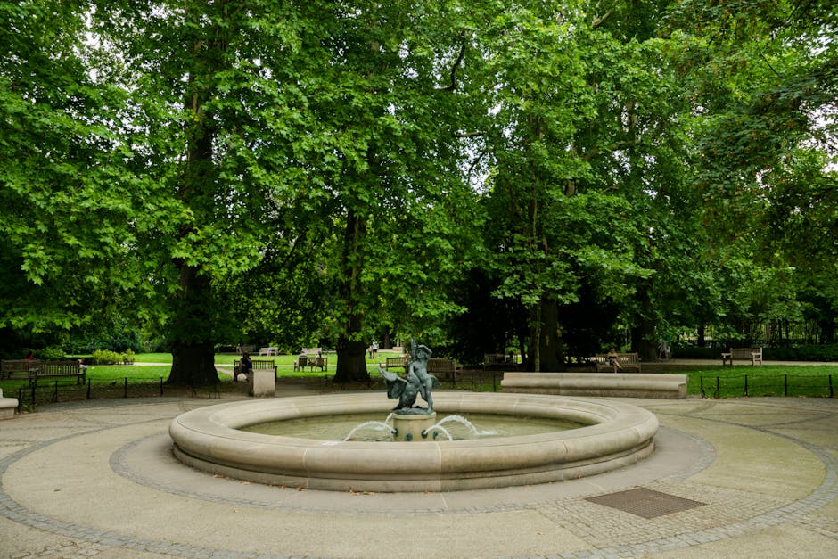 The image depicts a spacious urban park with a large open grassy area surrounded by mature trees with green and dark purple foliage. Several people are seated on benches along the periphery, some appear to be resting or relaxing. In the foreground, the branches of a tree extend into the view, framing the scene. The sky above is cloudy with patches of sunlight filtering through, creating a soft light across the landscape. The park's environment suggests a quiet, well-maintained space suitable for leisure activities. This setting could be part of a home relocation process, where furniture and boxes are transported into or out of nearby properties, possibly involving the use of a man and van service like Man and Van Ravenscourt Park, who handle residential removals and furniture transport within the area.