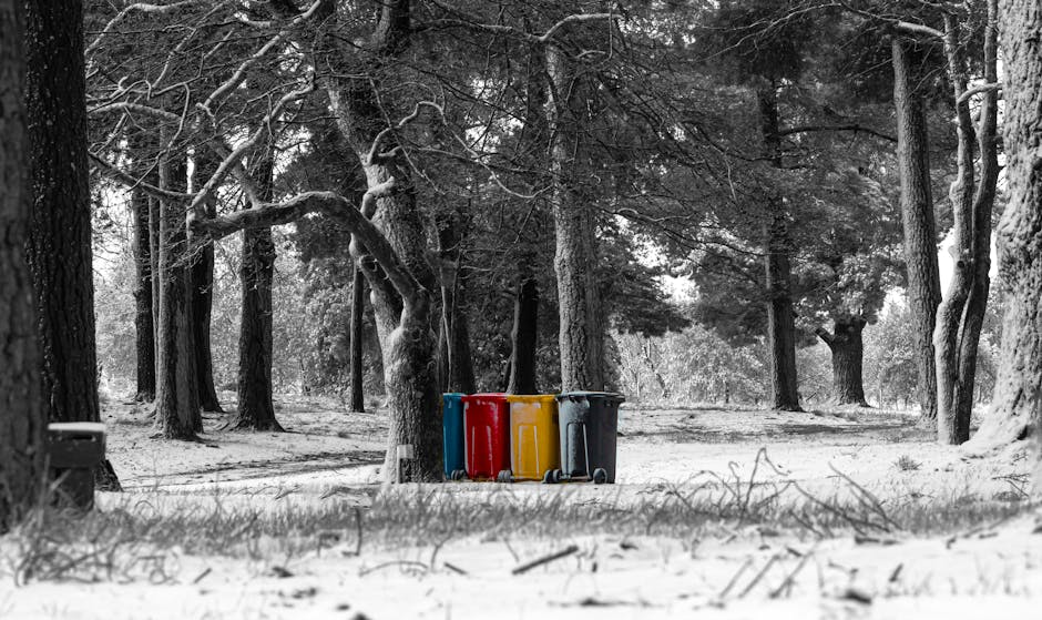 A person wearing a traditional conical hat and dark clothing with reflective strips is pushing a green wheelie bin along a sidewalk lined with trees, outstretched branches overhead casting shadows on