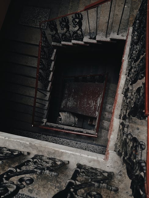 A narrow staircase inside a residential building with a wooden handrail and white balustrades, leading up from a patterned tiled floor area. At the top of the stairs, there is a windowsill with severa